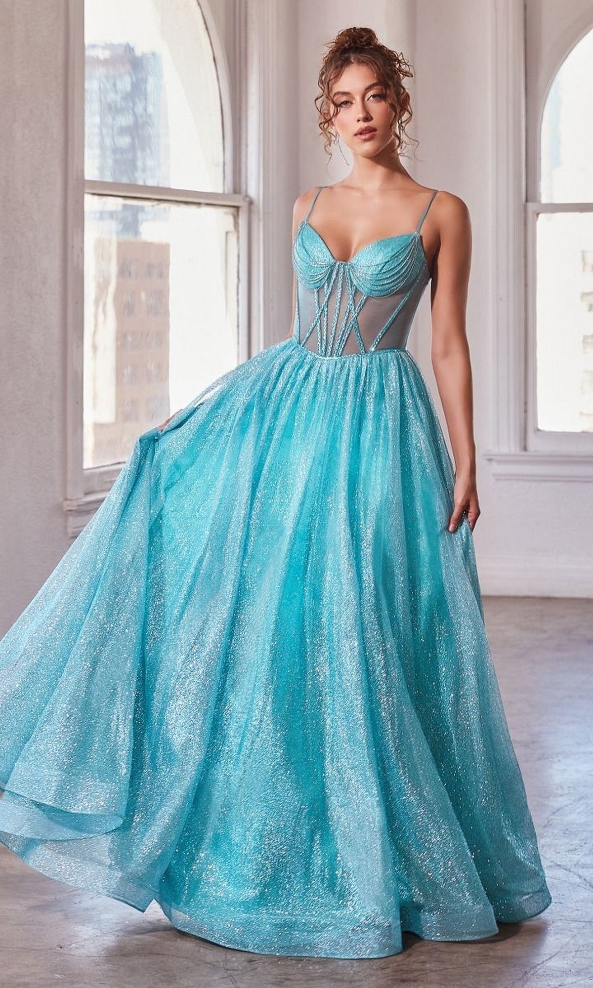 A woman stands in a sunlit room wearing the Sheer-Bodice Long A-Line Shimmer Prom Dress CD832—a sparkly blue formal gown with a fitted bodice and sheer panels—holding out its skirt.